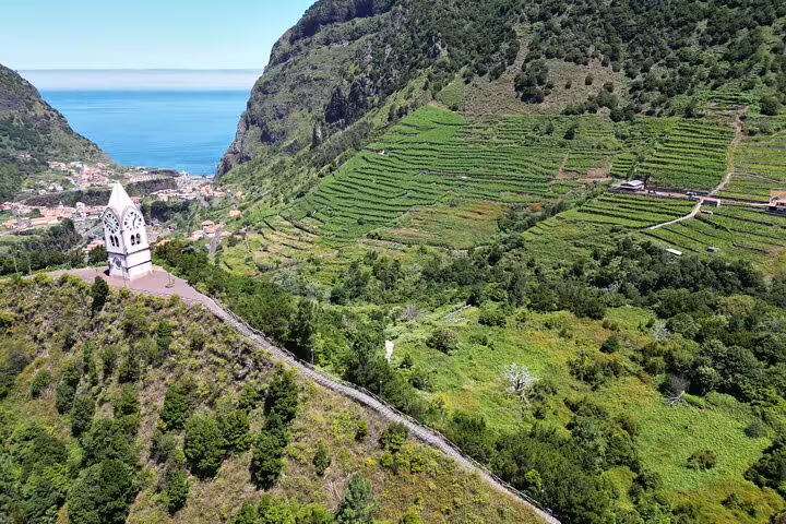 Scenic view of lush green terraced hills and a historic church overlooking the ocean on a 4x4 tour to Porto Moniz and Fanal.