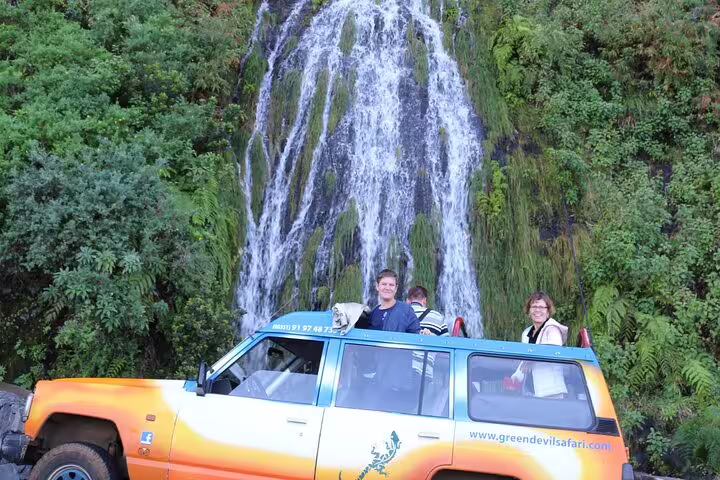 Tourists enjoy a scenic 4x4 adventure in Madeira's lush northwest, with an open-roof vehicle and stunning waterfall backdrop.