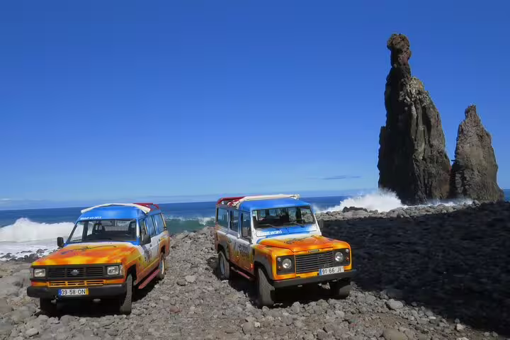 Two colorful 4x4 vehicles parked on a rocky beach in Madeira, with dramatic sea stacks and ocean waves in the background.