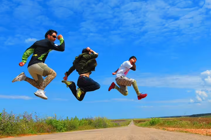 Adventurers leap joyfully on a sunny road during a 4x4 West Coast tour in the Natural Park, under a vibrant blue sky.