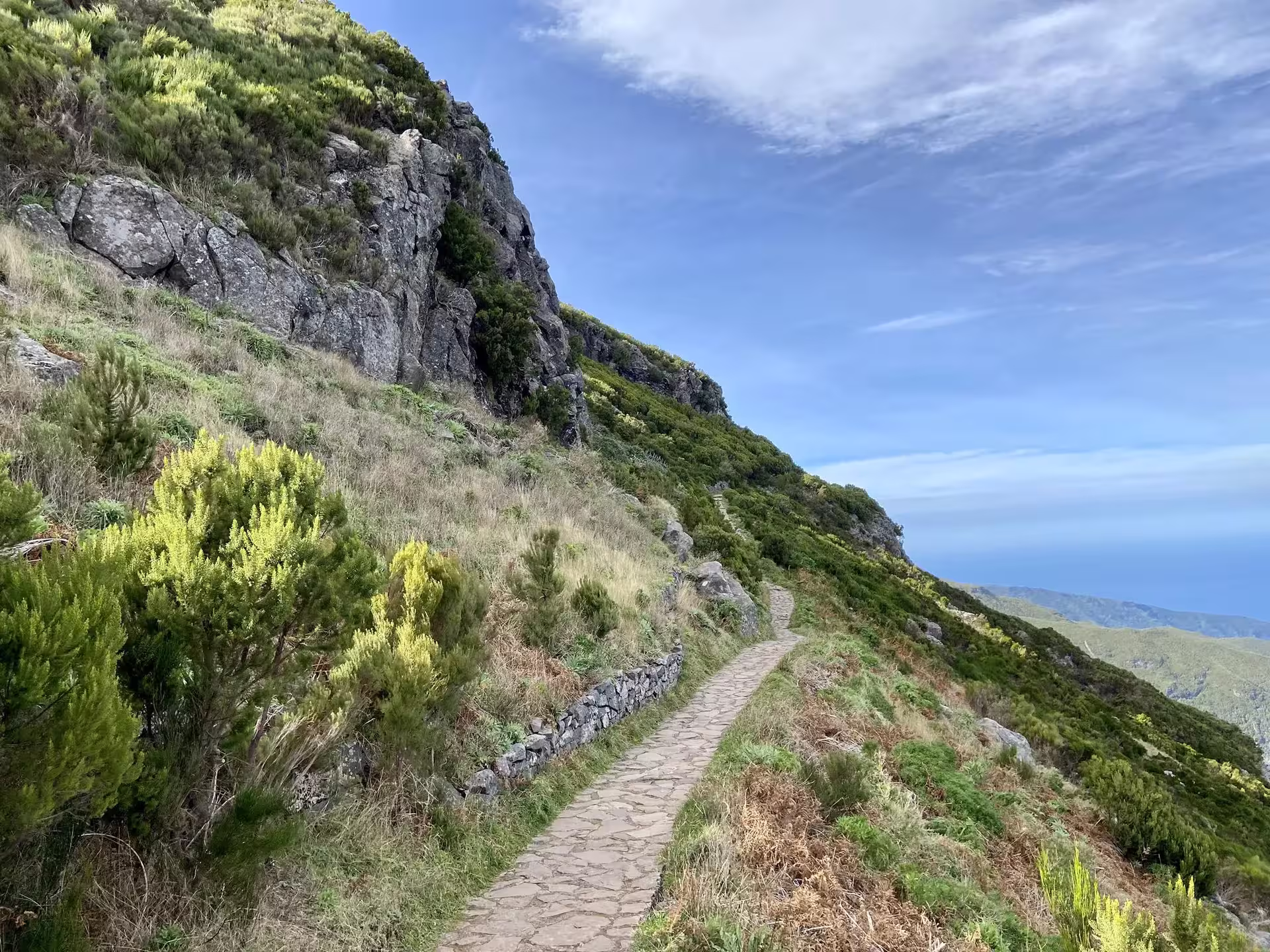 Narrow stone trail along steep hillside offering panoramic views of green landscape on Above the Skies' Hike.