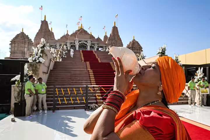 A woman in traditional attire blows a conch shell at the entrance of a grand Hindu temple in Abu Dhabi under a clear sky.