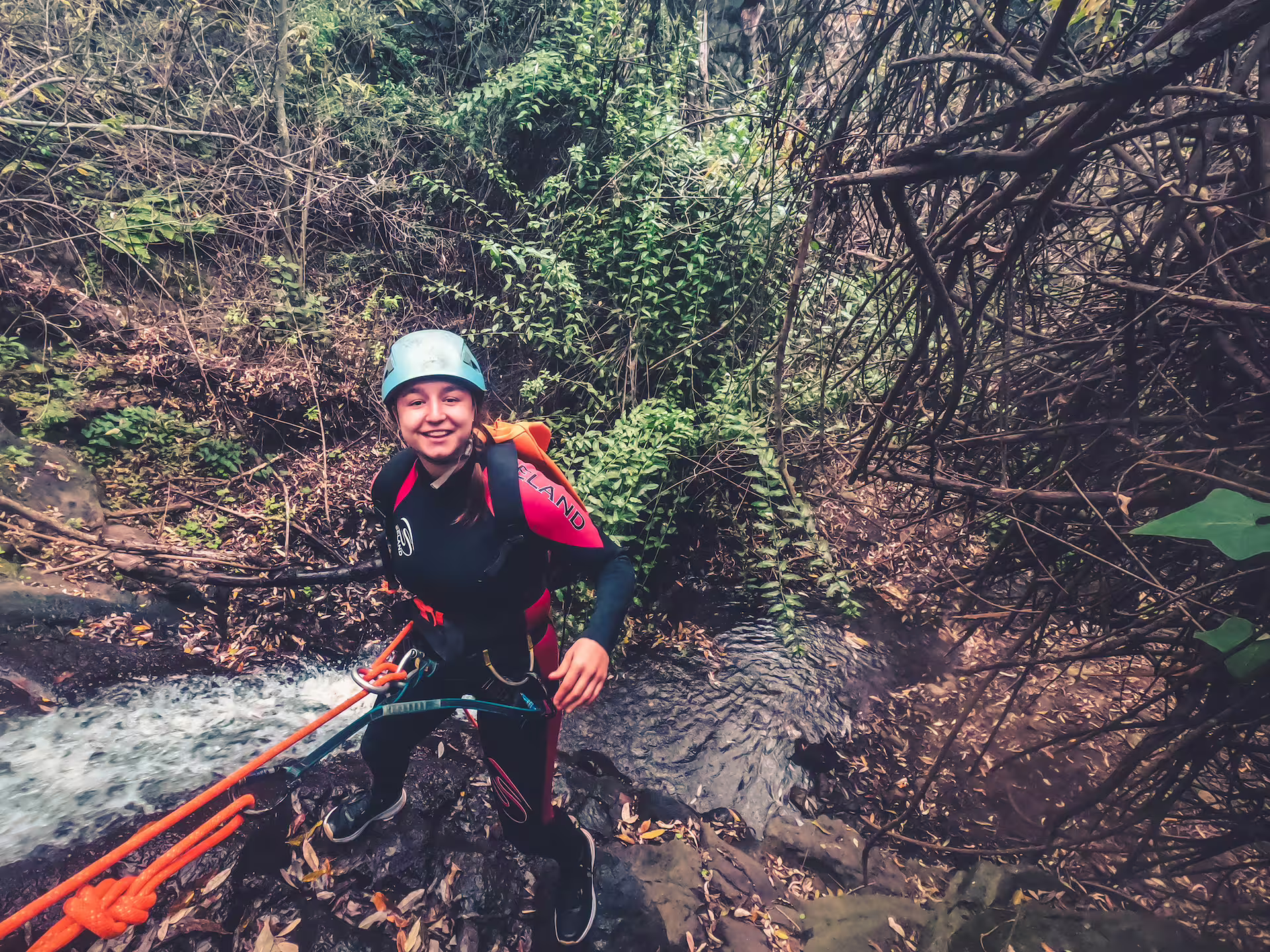 Adventurer joyfully canyoning through lush rainforest, equipped with harness and helmet for a thrilling outdoor experience.