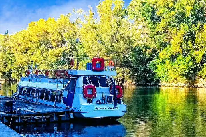 Tour boat Agrippina Maggiore moored on the Tiber River amid green trees for Ostia Antica cruise and archaeological day trip from Rome