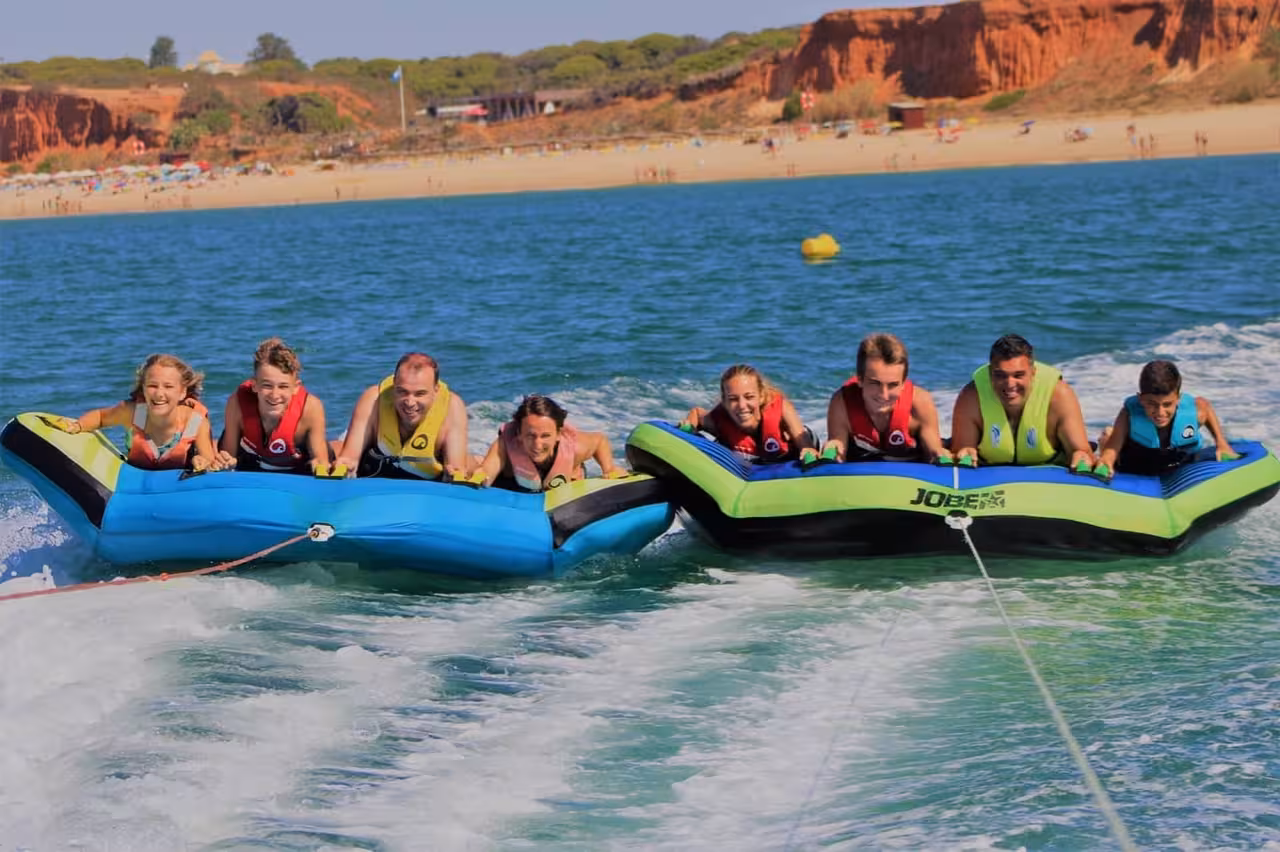 Group of friends riding inflatable Airstream tubes across turquoise ocean waves near a sunny Algarve beach