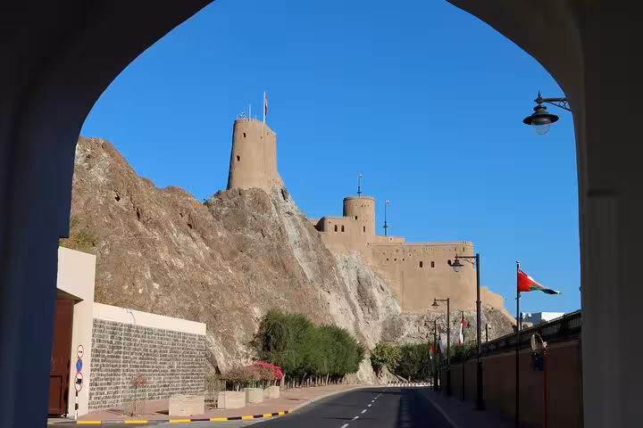 View of historic Al Jalali Fort on rocky cliffs above a quiet road lined with Omani flags on a Half-Day Muscat City tour