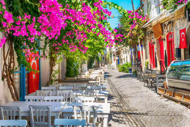 Narrow cobblestone street in Alaçatı with rows of white tables and chairs under vibrant pink flowers and Turkish flags.