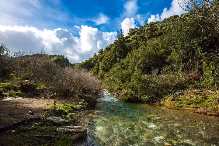 Crystal-clear stream flowing through lush green valley on Ikaria Hike: Alama Spring, under dramatic Aegean island skies