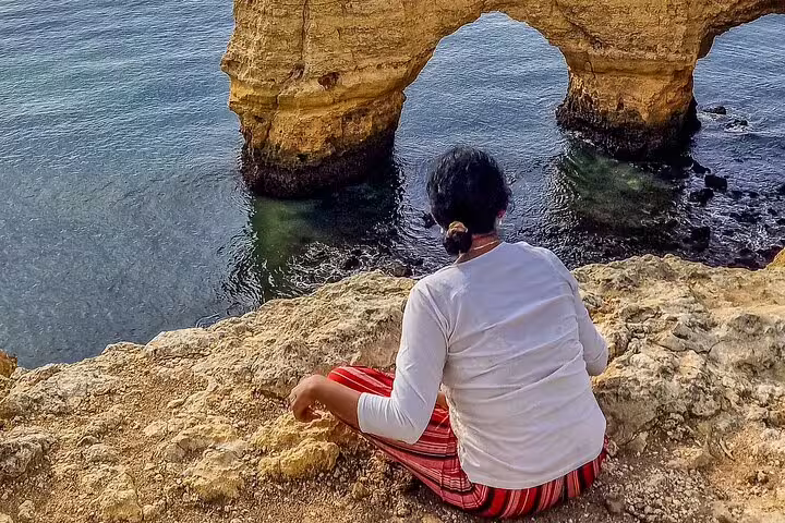 Person sitting on rocky cliff overlooking stunning Algarve coastline and Benagil Caves during private tour from Lisbon.
