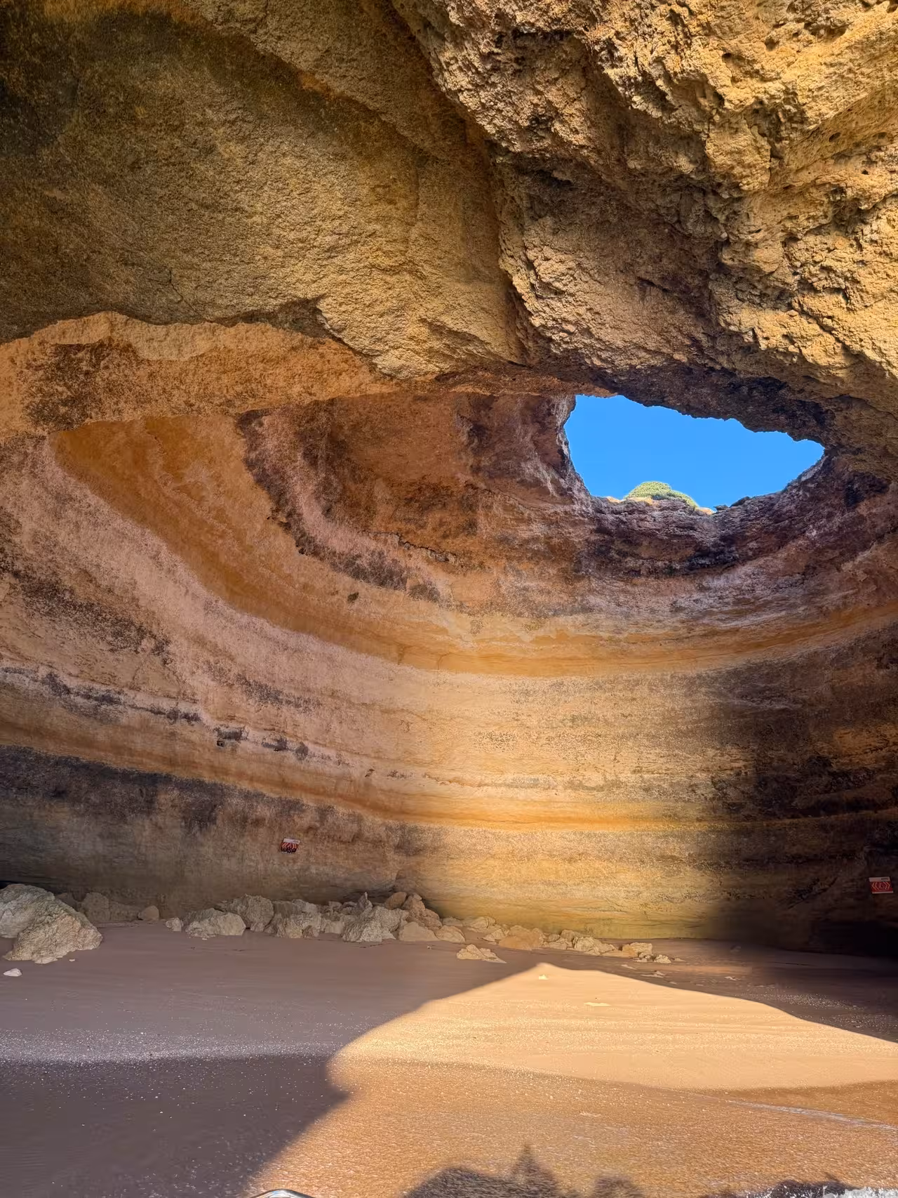 Sunlit sand and rock formations inside a vast Algarve sea cave with natural skylight opening to blue sky above
