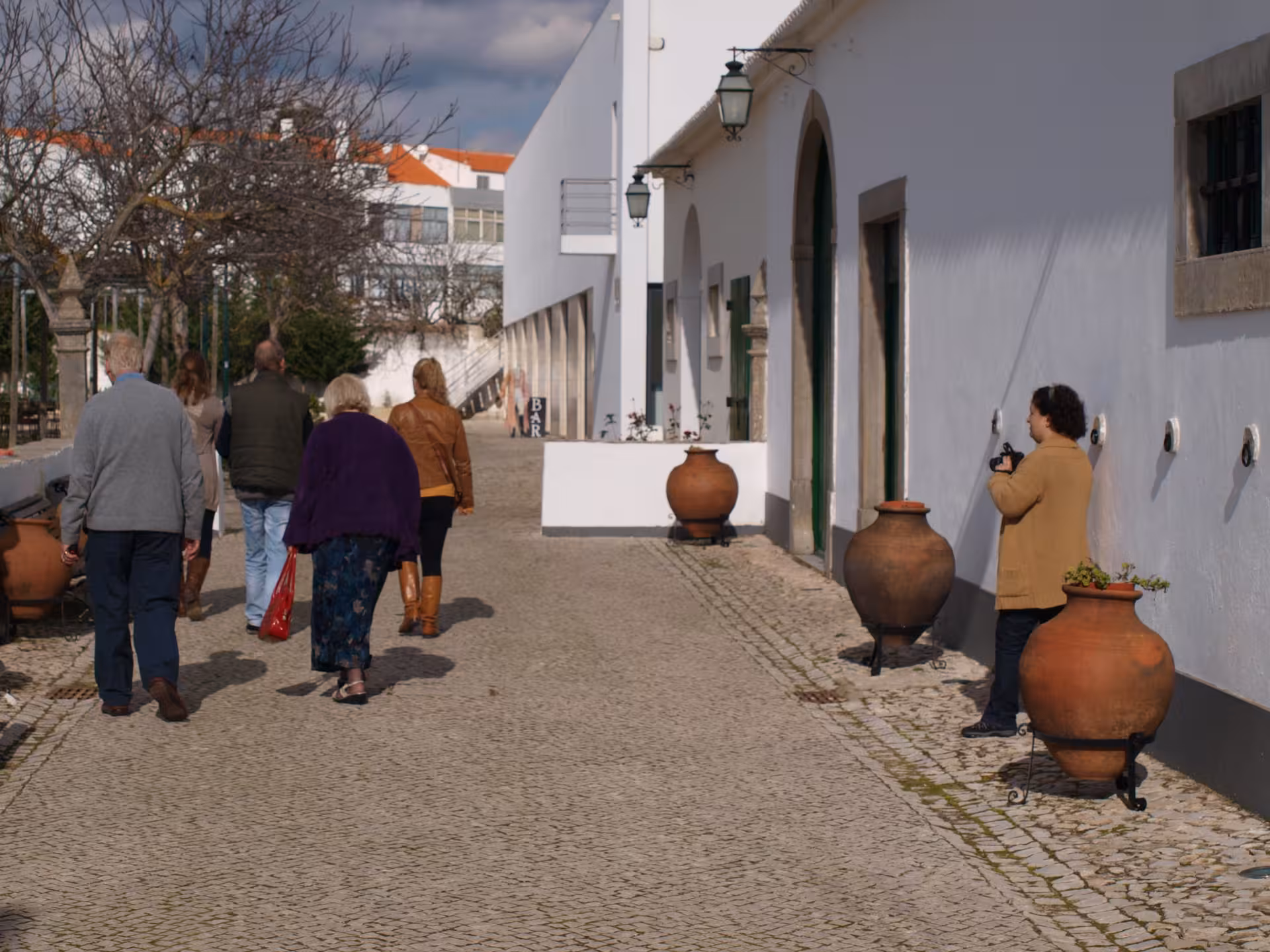 Visitors strolling a cobbled street past whitewashed houses and terracotta jars in a historic Algarve village