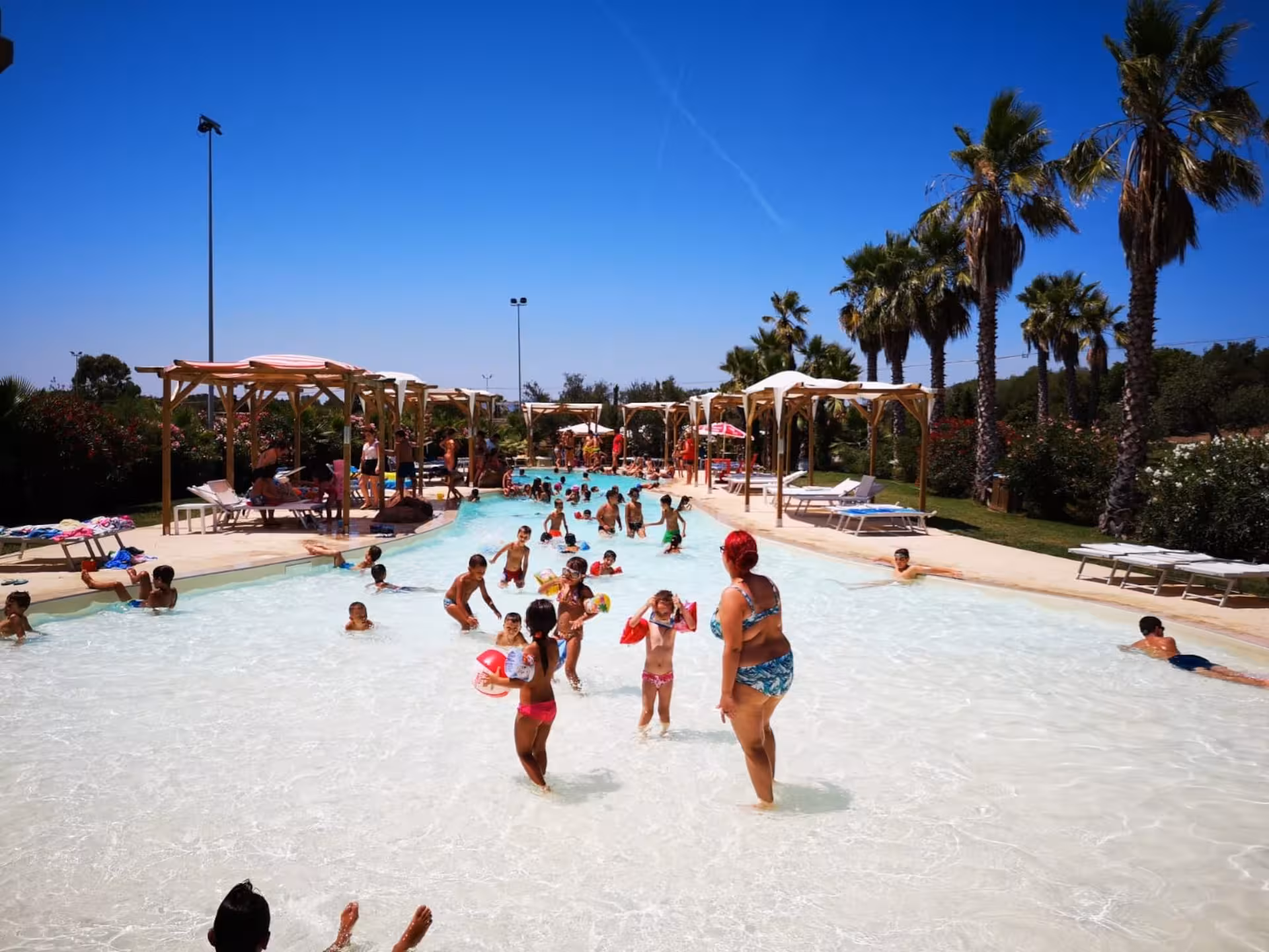 Families enjoy a sunny day at Alghero Butterfly House pool, surrounded by palm trees and vibrant flowers.