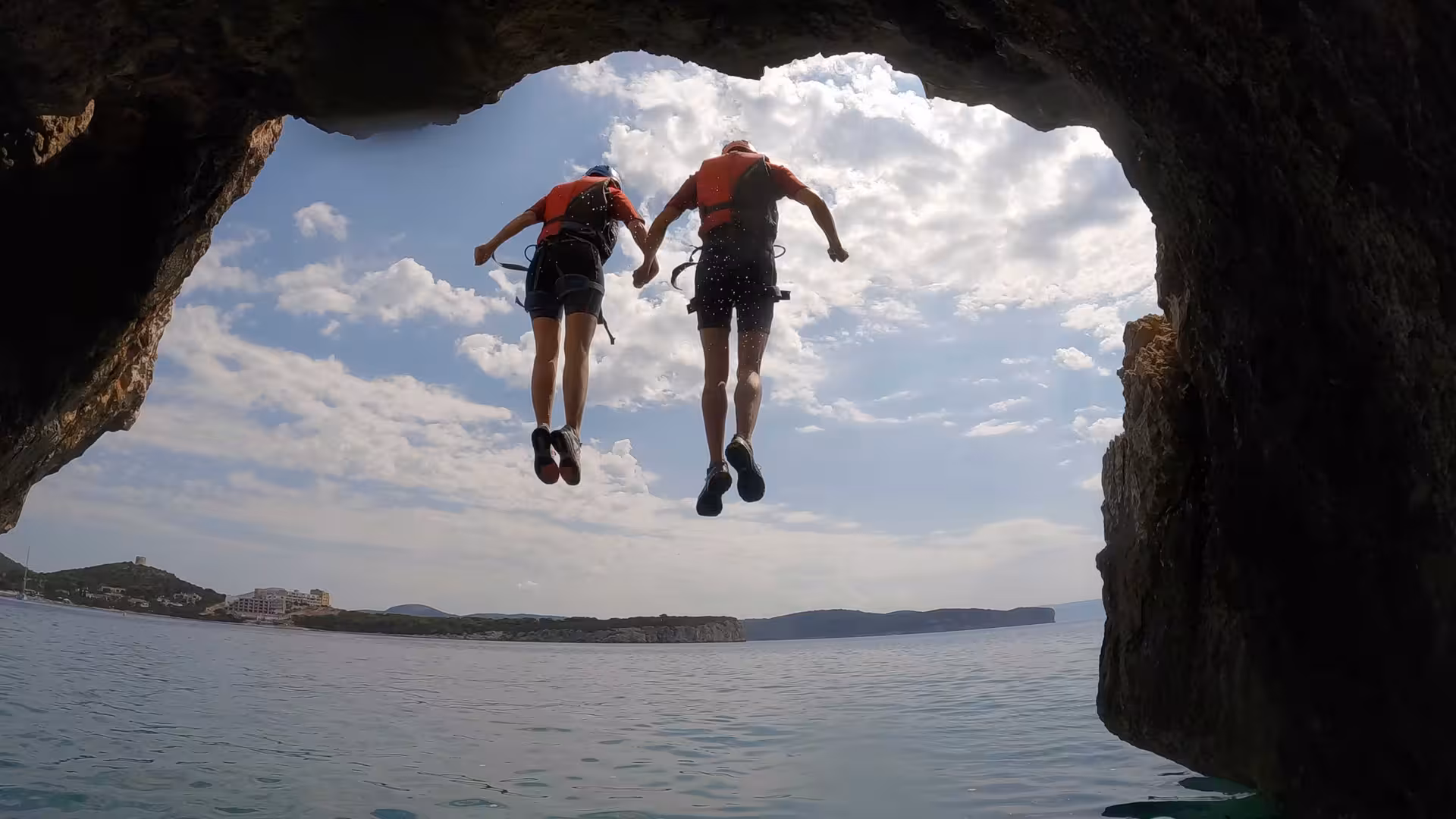 Pair of thrill-seekers leap from a rocky arch into the sea at Cala Dragunara, Alghero, enjoying coasteering fun.