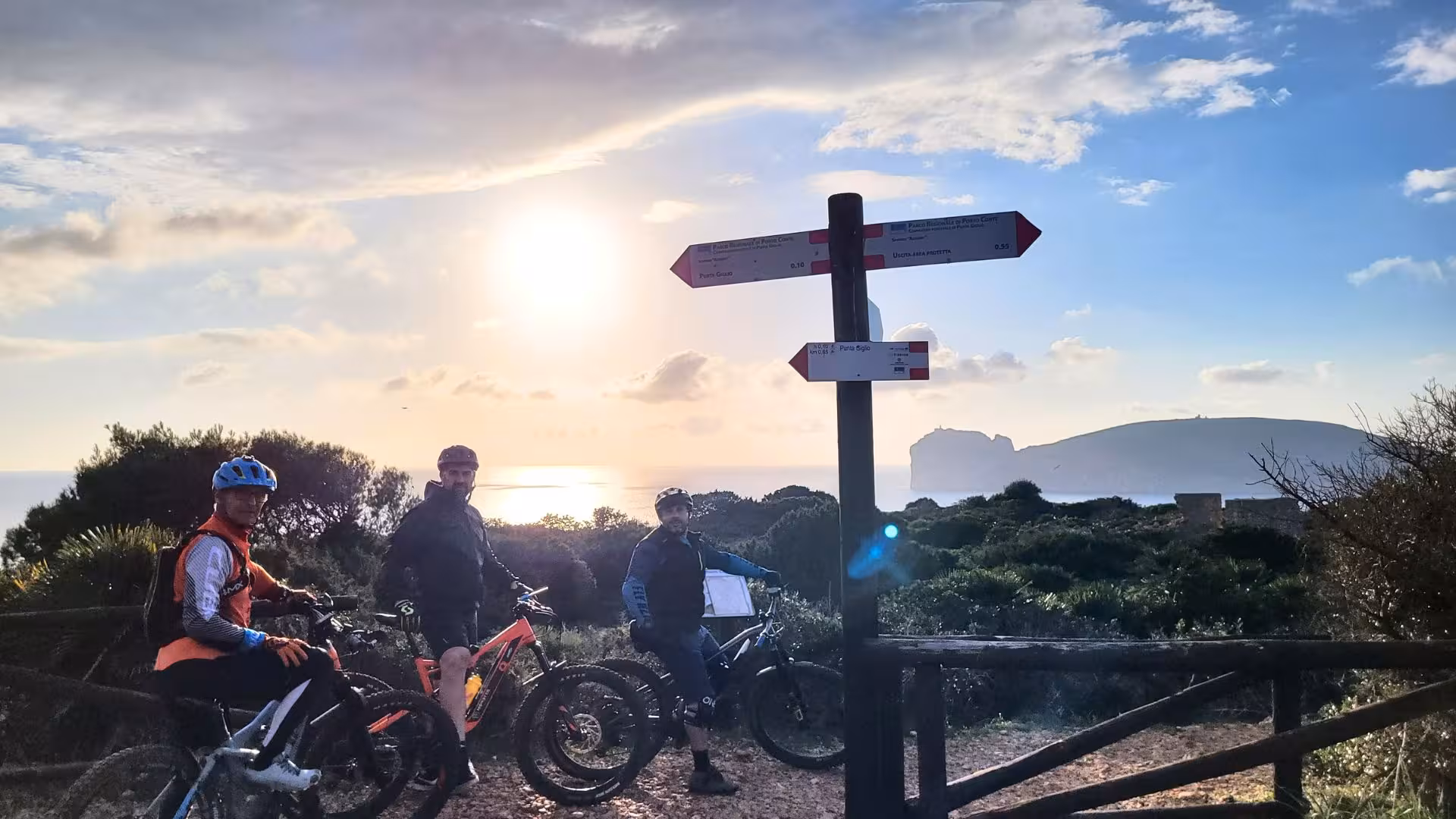 Cyclists on ebike tour at Capo Caccia in Alghero with scenic sunset view and signpost.