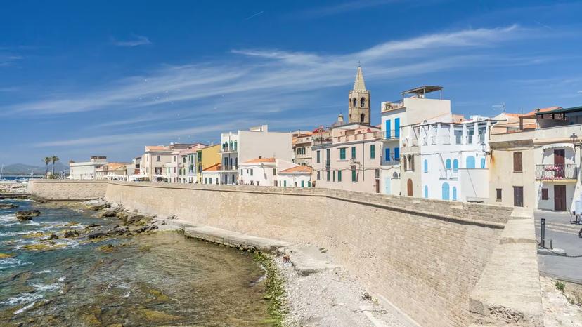 Picturesque coastal view of Alghero's historic buildings along the city walls under clear blue skies.