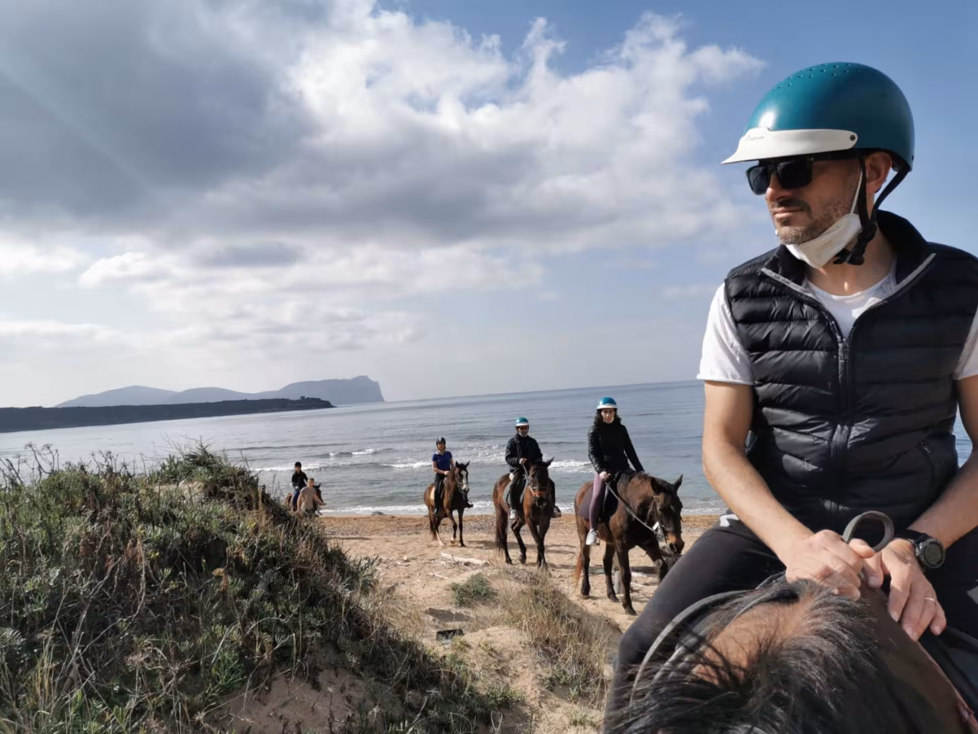 Horseback riders traverse a sandy path by the sea in Alghero, with stunning coastal views and lush greenery.