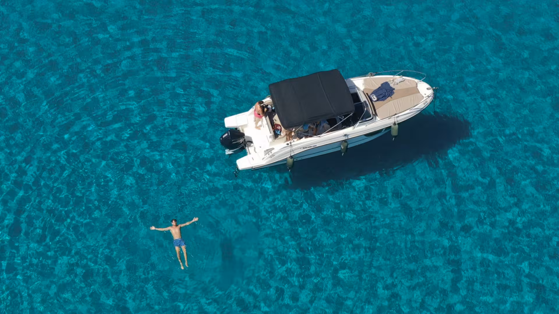 Aerial view of a motor boat and a person swimming in crystal clear waters on a half-day Alghero boat tour.