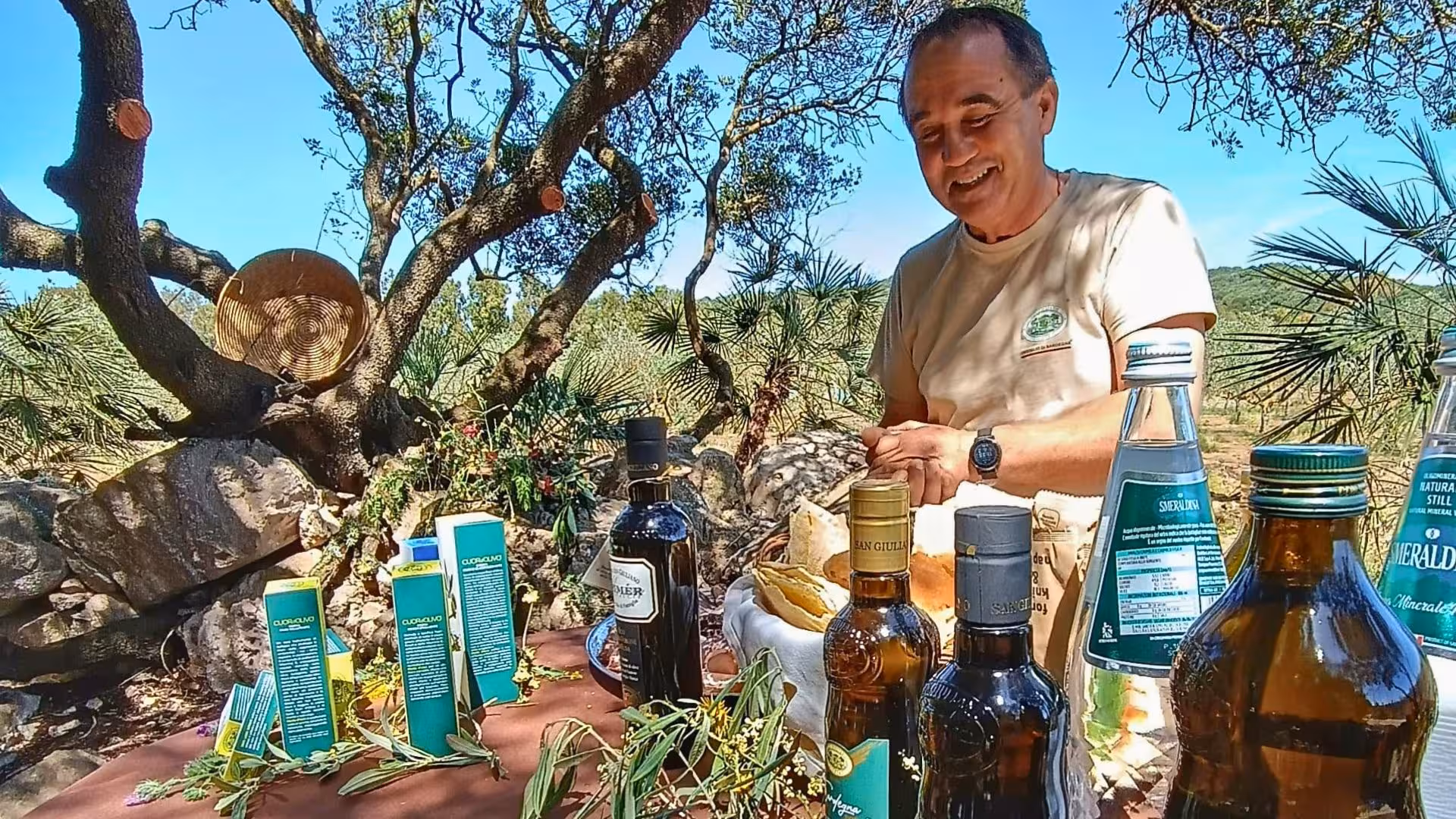 Smiling guide presents olive oil bottles and products under an olive tree during Alghero grove tour with tasting.