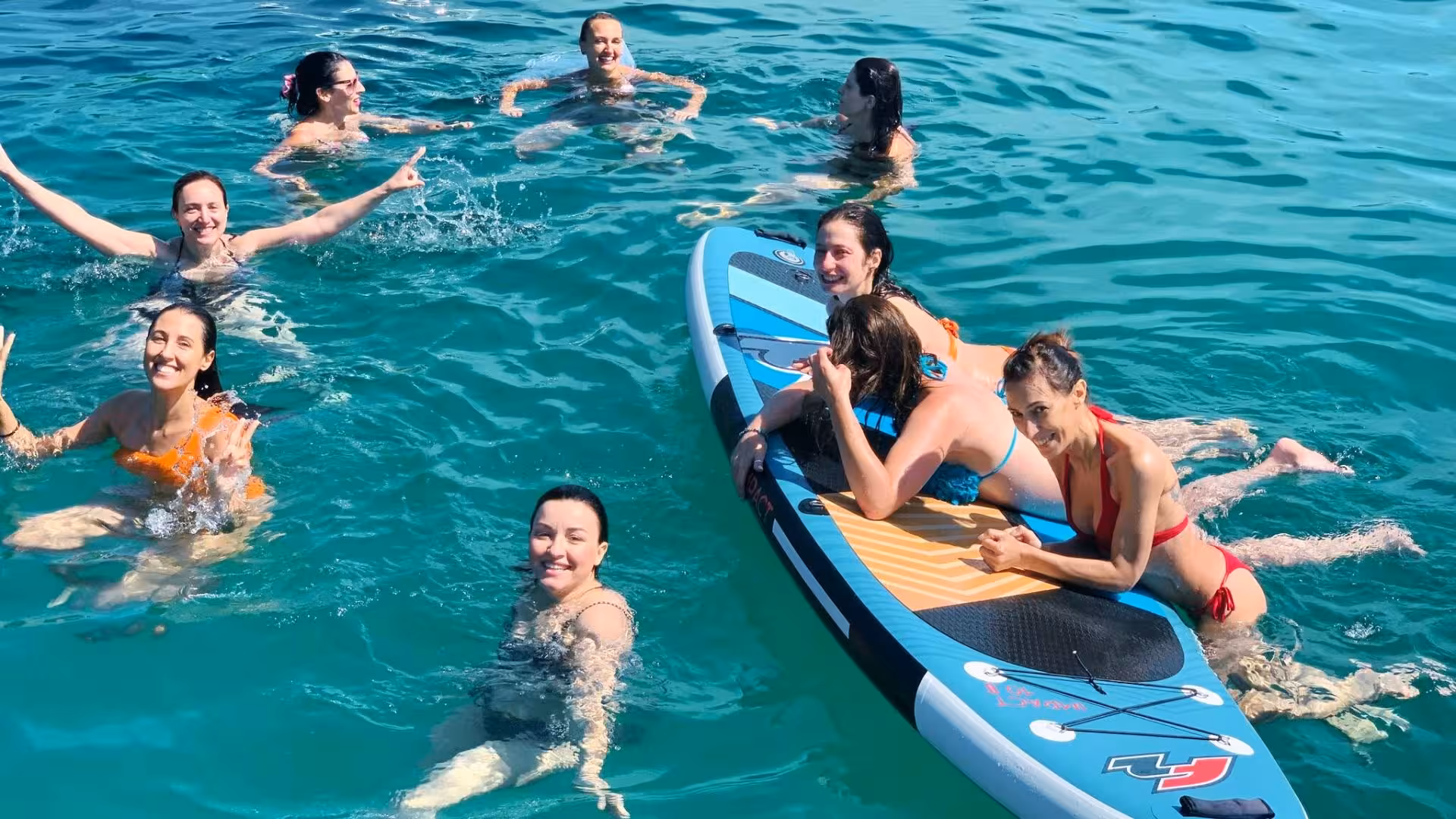 Tour group swimming and relaxing on a paddleboard in the clear waters of Porto Conte Park during a sailing tour.