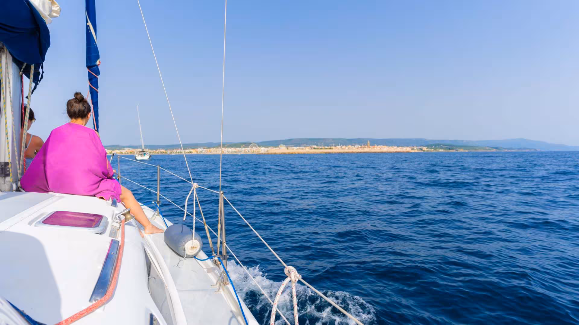 Tourists relaxing on a sailboat with a view of Alghero's coastline during a Porto Conte Park sailing tour.