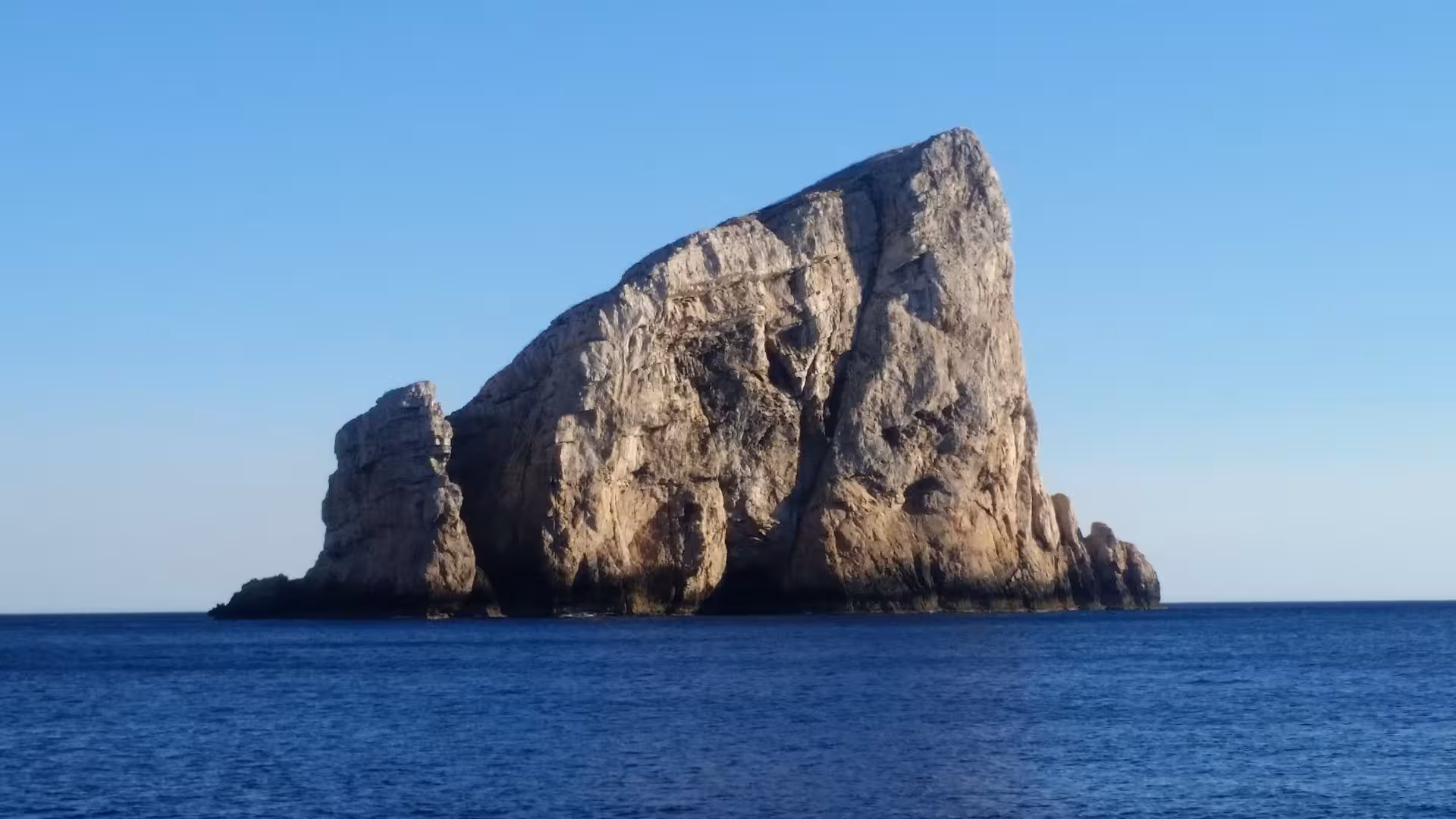 Impressive rock formation emerges from the clear blue sea on a serene Alghero sailboat excursion.