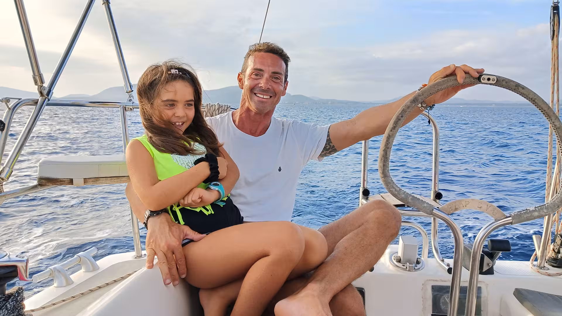 Father and daughter smile joyfully on a sailing boat in Alghero, with picturesque ocean views in the background.