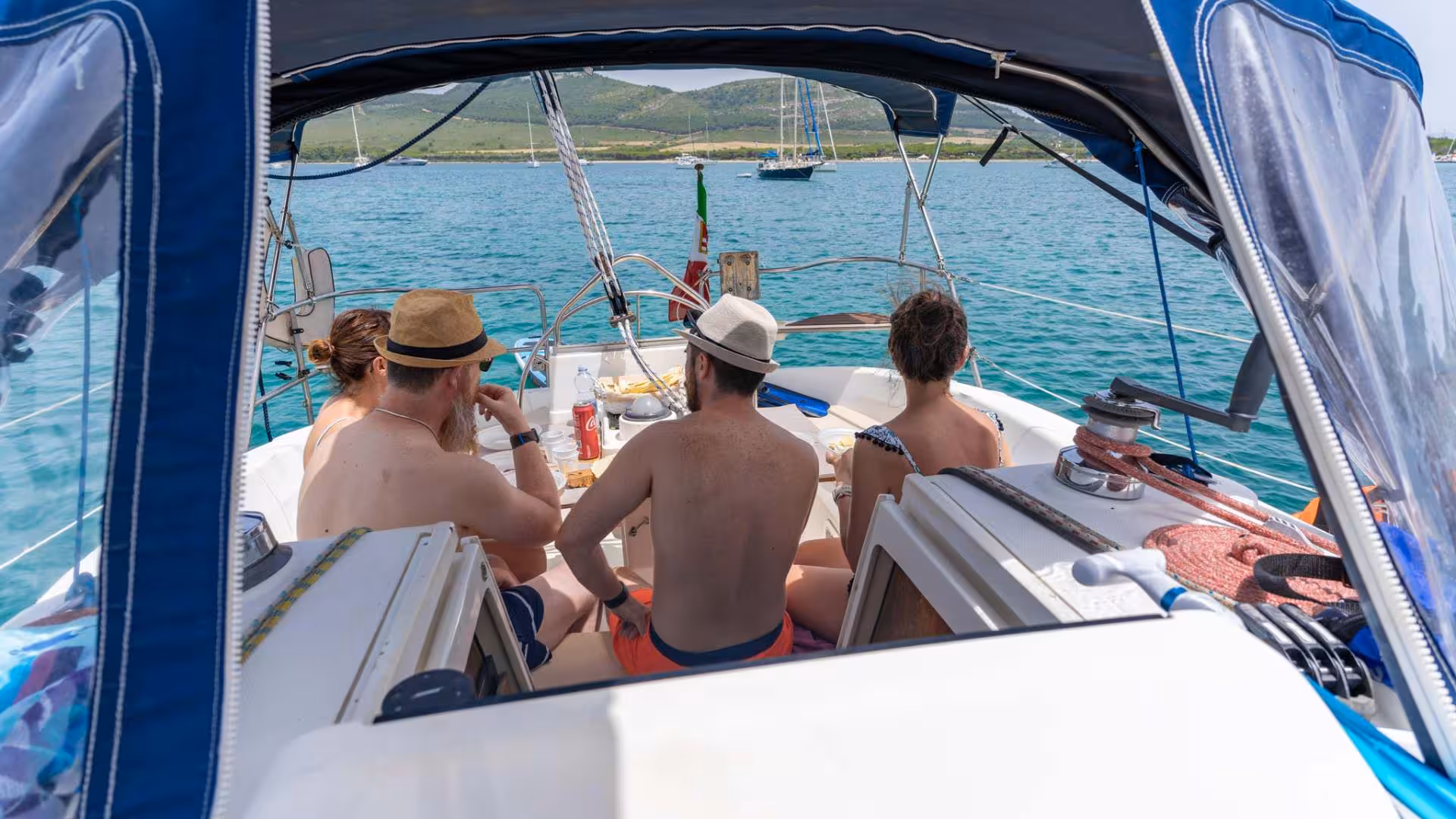 Group enjoying a meal on a sailboat tour in Porto Conte Park, Alghero, surrounded by clear blue waters.