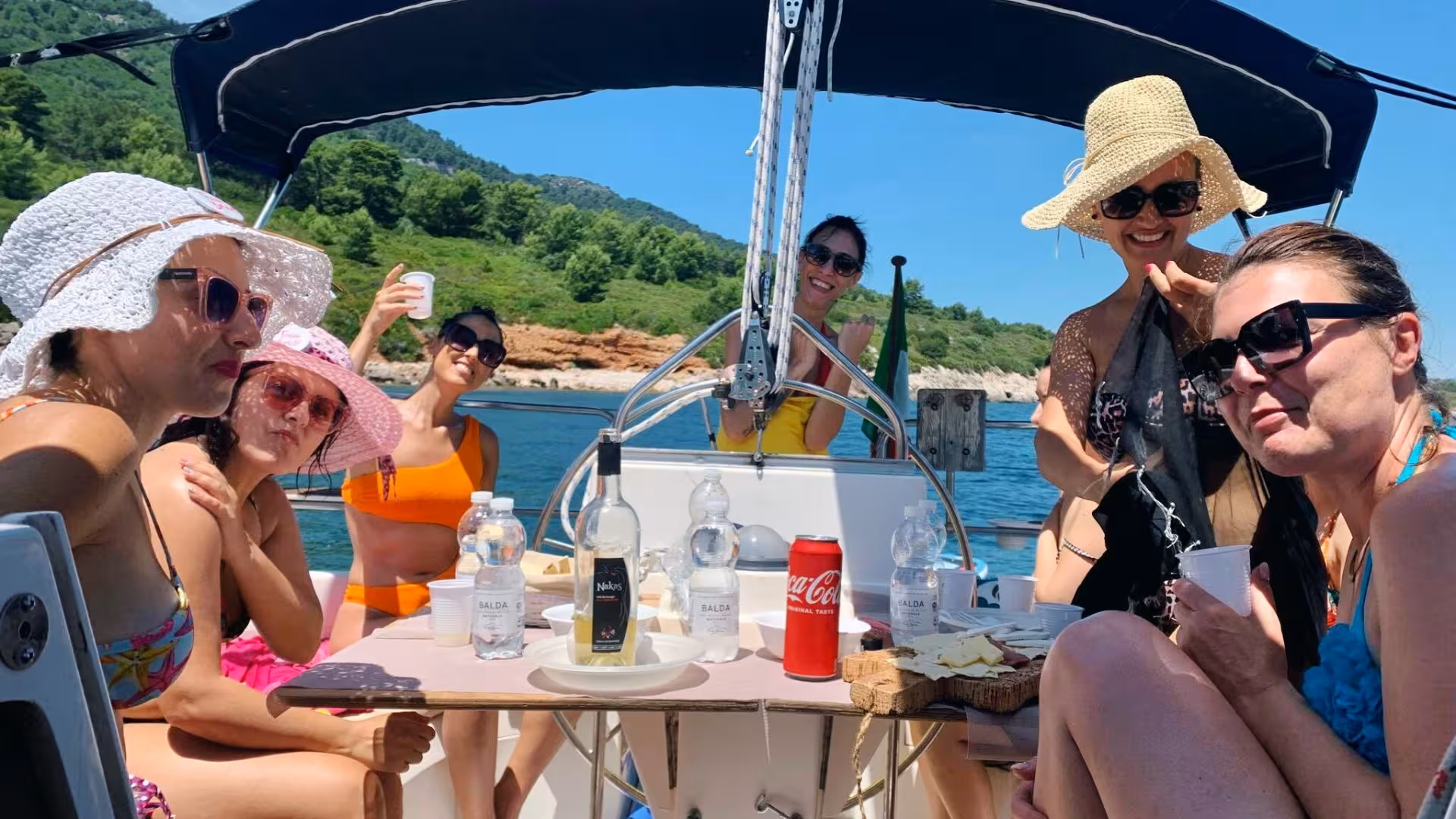 Group of friends enjoying drinks and snacks on a sailing tour in Porto Conte Park, surrounded by scenic landscapes.