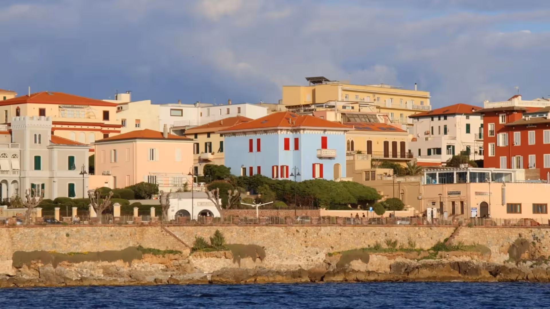 Colorful buildings along Alghero's coastline, viewed from the sea during a sailing tour at sunset.