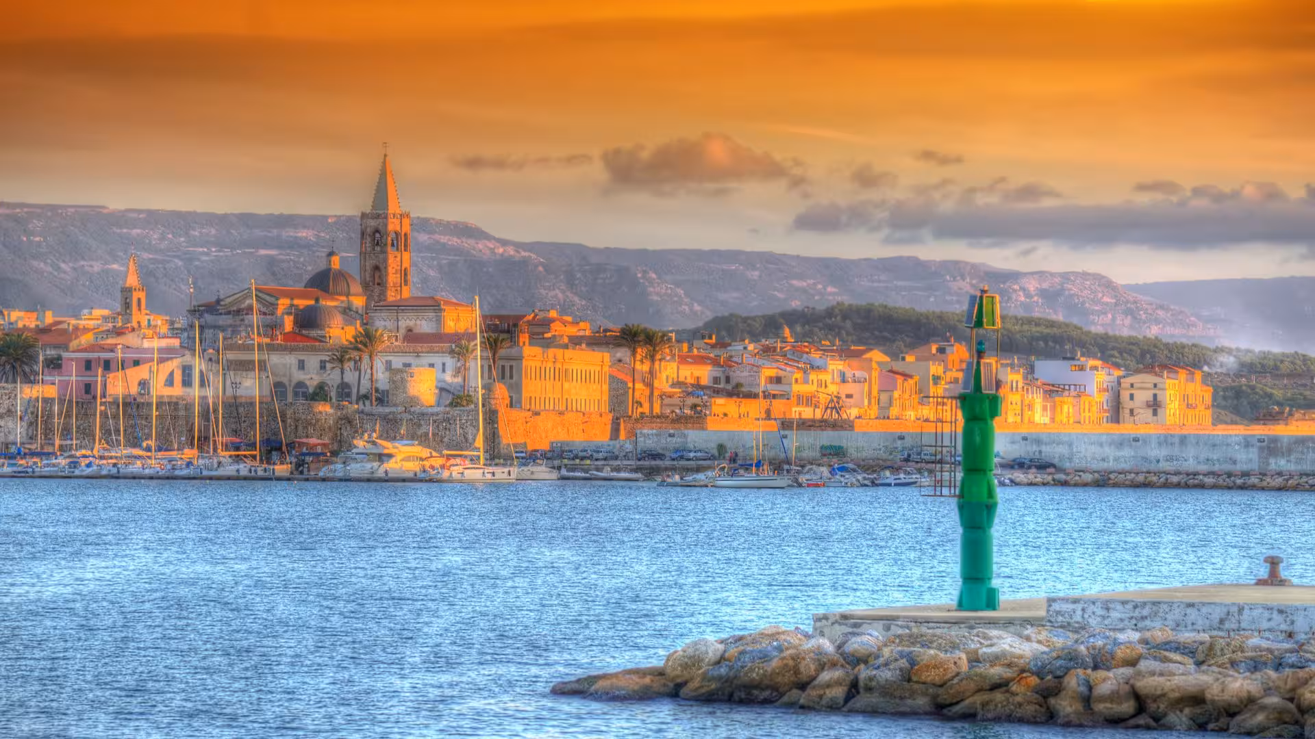 Scenic view of Alghero's historic skyline and harbor under a vibrant sunset sky on a sailing excursion.