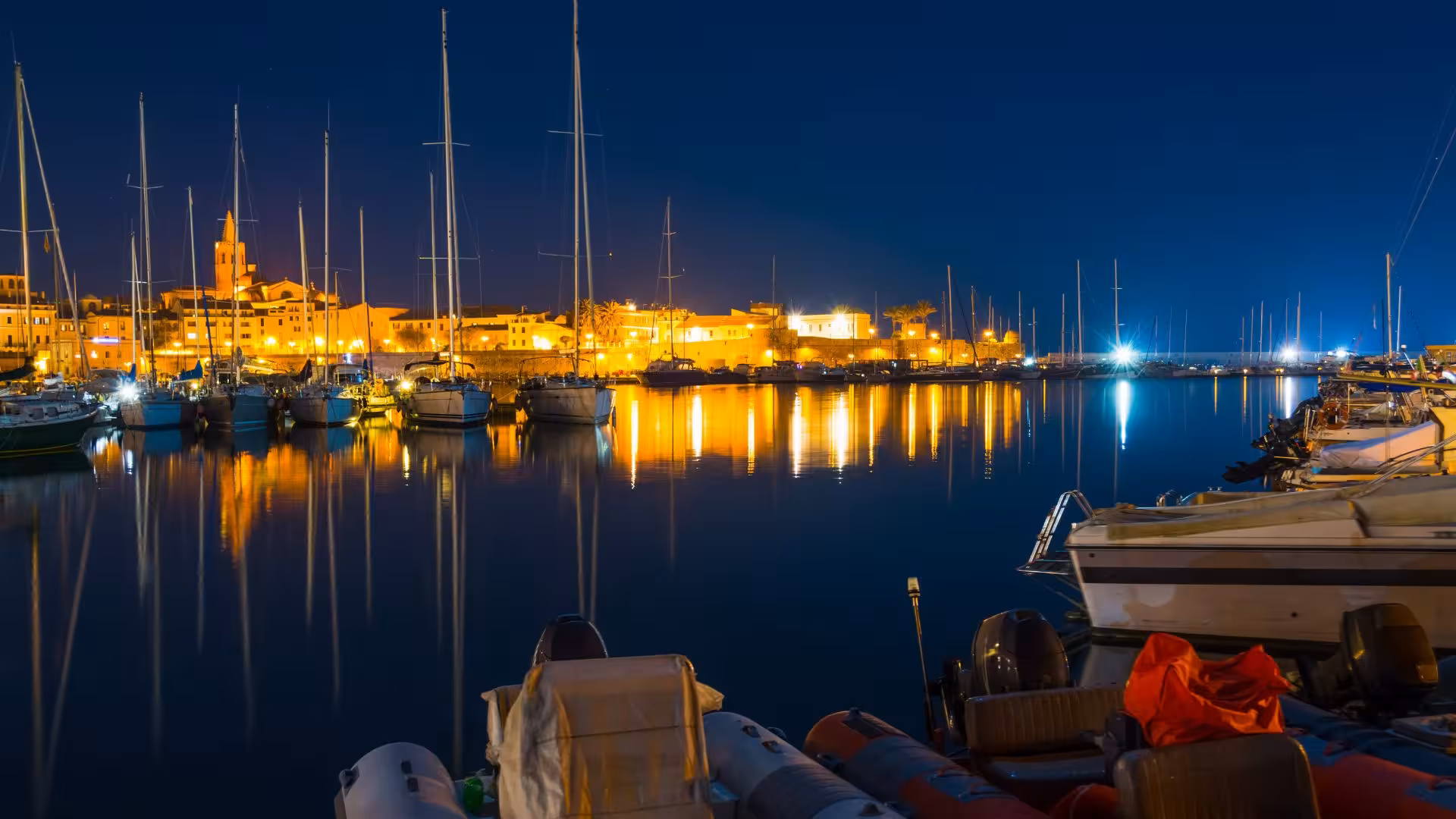 Night view of Alghero harbor with illuminated boats reflecting on the calm water, ideal for a sunset sailing tour.