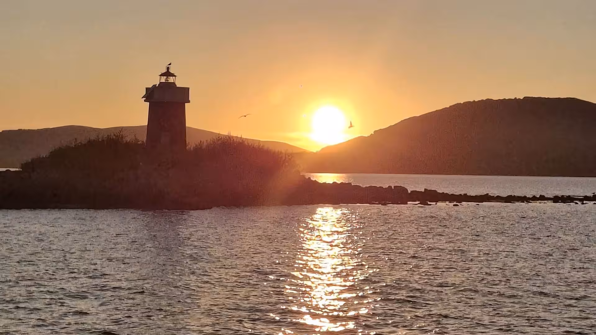 Golden sunset behind a lighthouse on a serene Alghero coastline, perfect for an evening sailing boat aperitif.