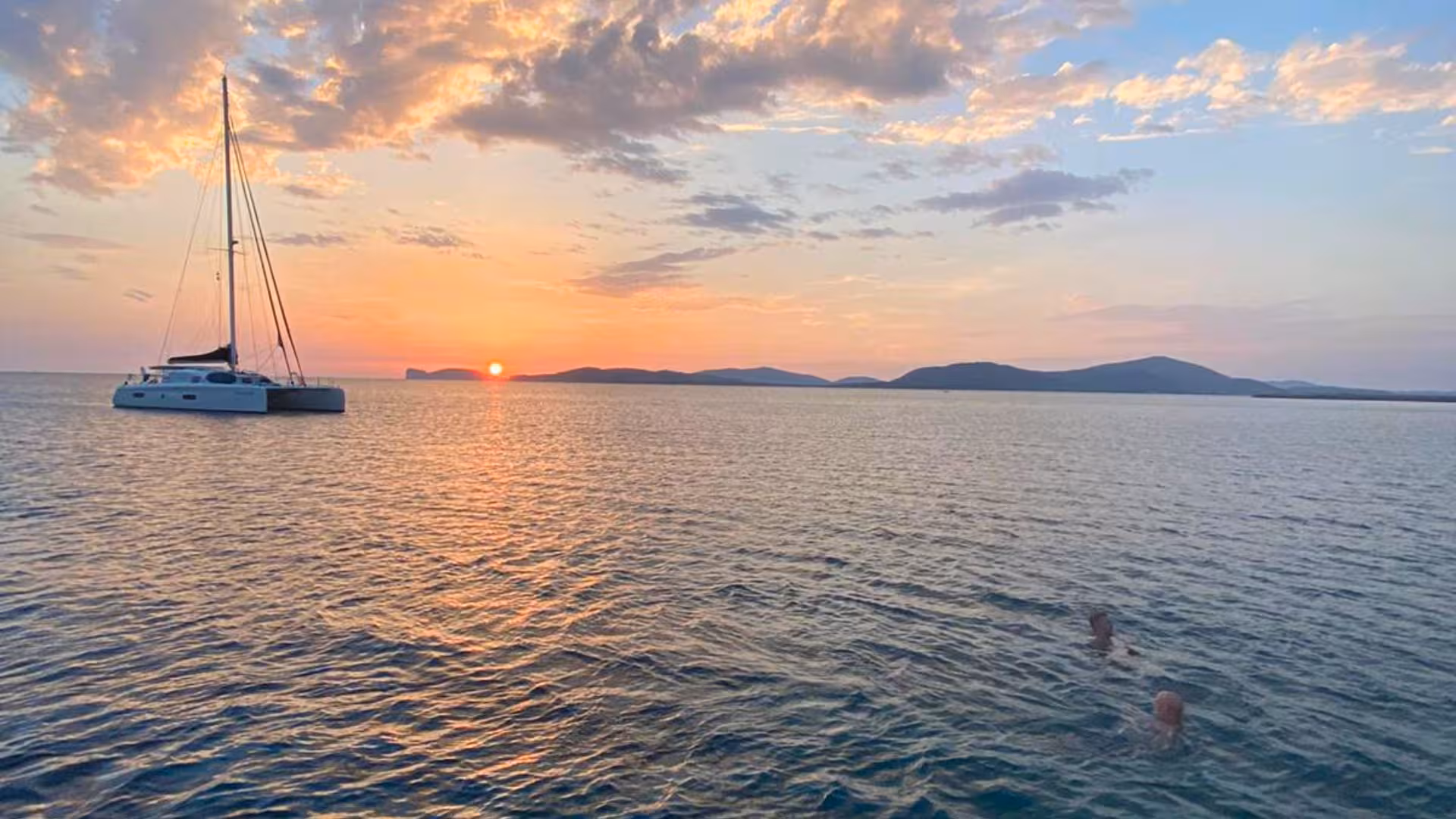 Catamaran anchored in Alghero waters at sunset, with swimmers enjoying the serene sea and vibrant sky.