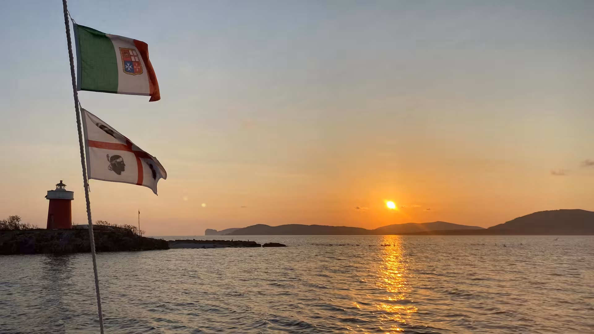 Scenic sunset over Alghero with lighthouse and flags on a sunset sailing tour with aperitif.