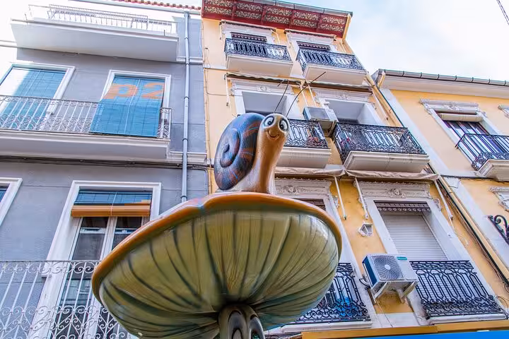Quirky snail sculpture on a mushroom in front of colorful, traditional Alicante buildings during the city walk.