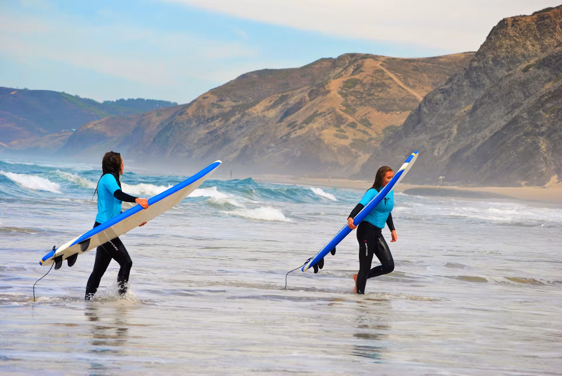 Two surfers in wetsuits carrying longboards into Aljezur beach waves during a guided full-day surf lesson tour