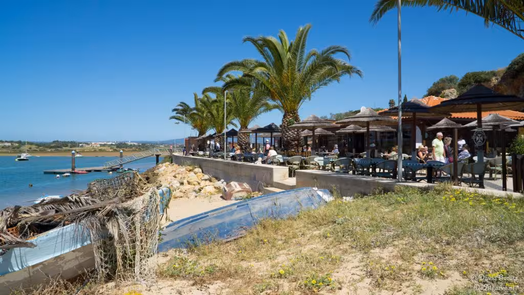 Riverside café terrace with palm trees, fishing boats and sandy shore near Alvor Estuary on Algarve cultural tour