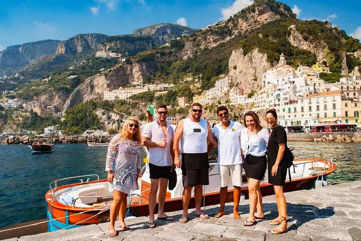 Tourists posing by a boat with stunning Amalfi Coast cliffs and charming towns in the background.