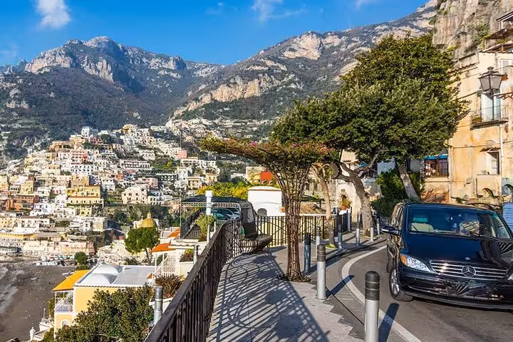 Scenic view of Positano along the Amalfi Coast with a minivan parked on a winding road under a clear blue sky.