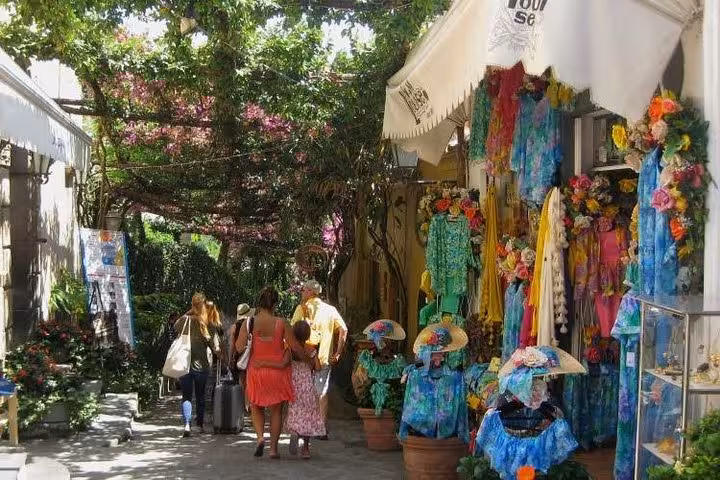 Tourists explore a charming Amalfi Coast alley lined with colorful shops and lush greenery on a sunny day.