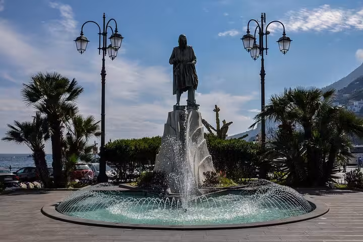 Statue and fountain in an Amalfi Coast plaza, surrounded by palm trees and overlooking the sea under a blue sky.
