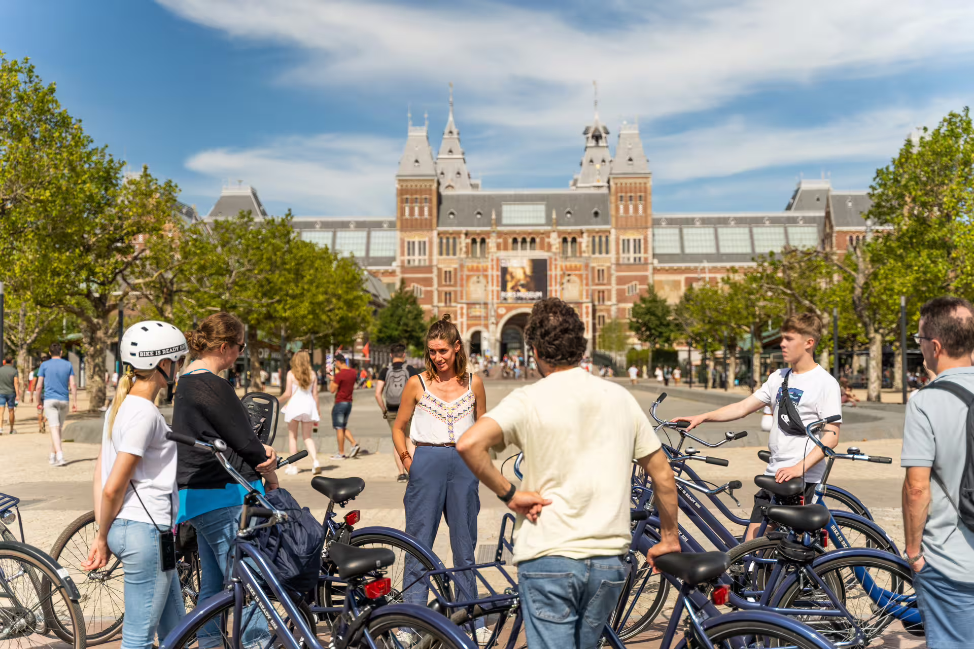 Group of tourists on bikes in front of Rijksmuseum in Amsterdam, enjoying a guided tour with canal cruise, cheese, and drinks.