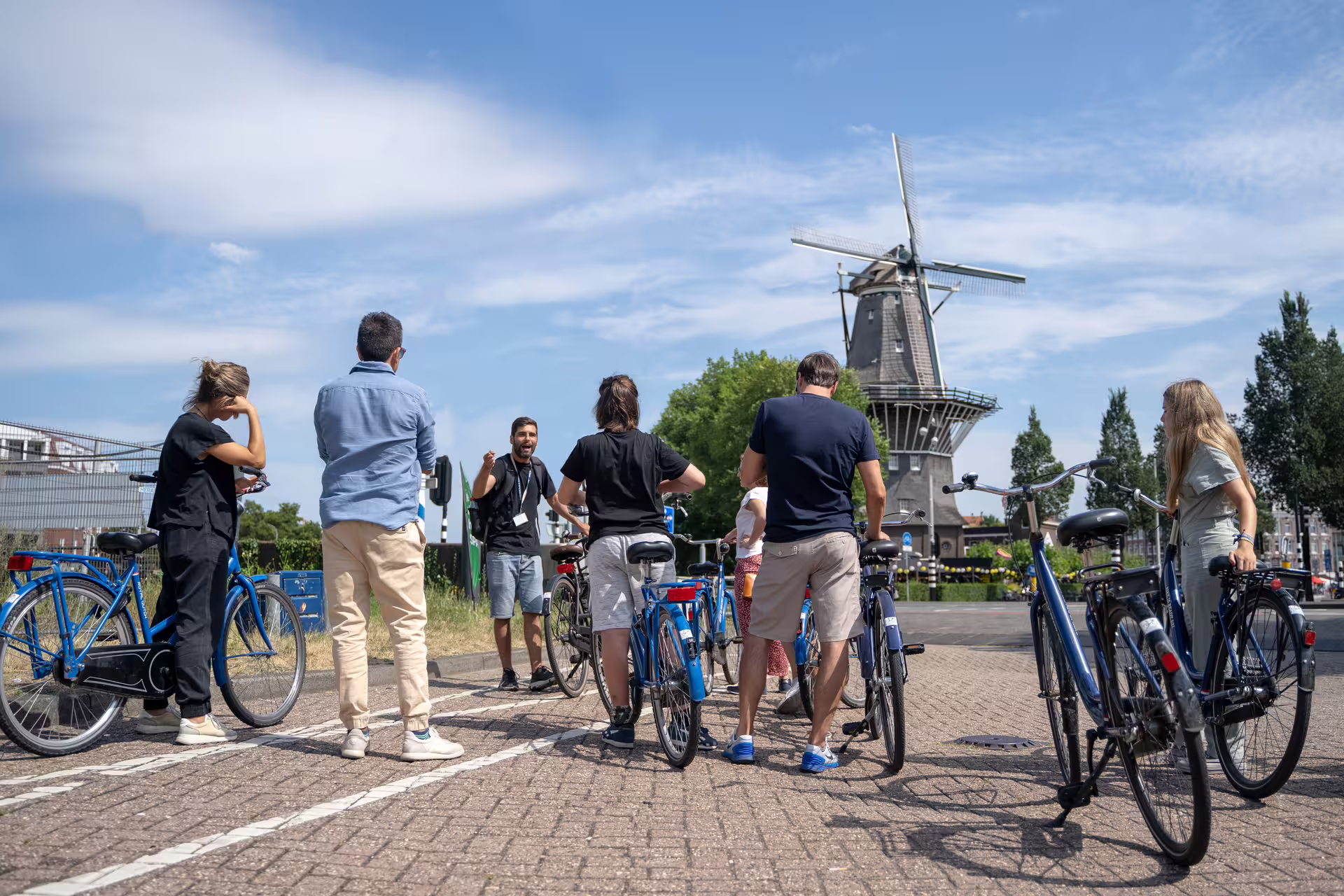 Cyclists gather near an iconic windmill in Amsterdam during a guided bike tour with included canal cruise, cheese, and drinks.