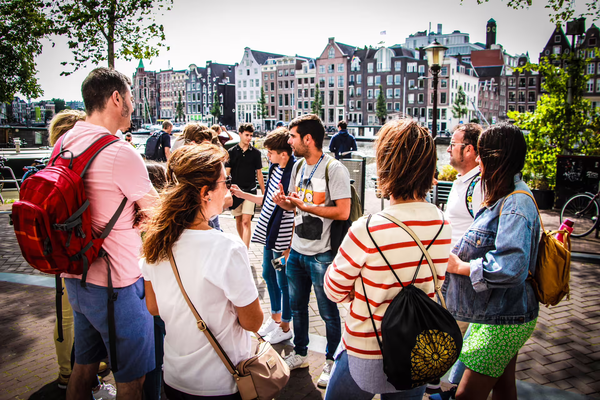 A group of tourists enjoys a guided walking tour in Amsterdam, with historic canal houses in the background on a sunny day.