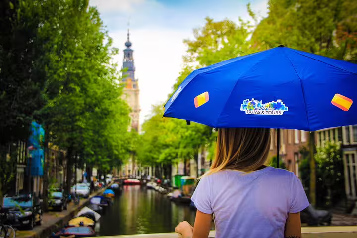A tourist with a blue umbrella overlooks an Amsterdam canal, highlighting the walking tour and cheese tasting experience.