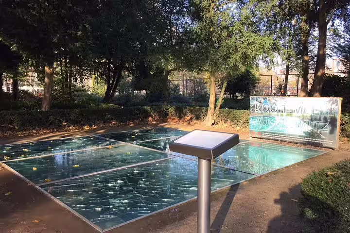 Reflective memorial glass monument in Amsterdam Jewish Quarter, surrounded by trees, part of private historical walking tour.