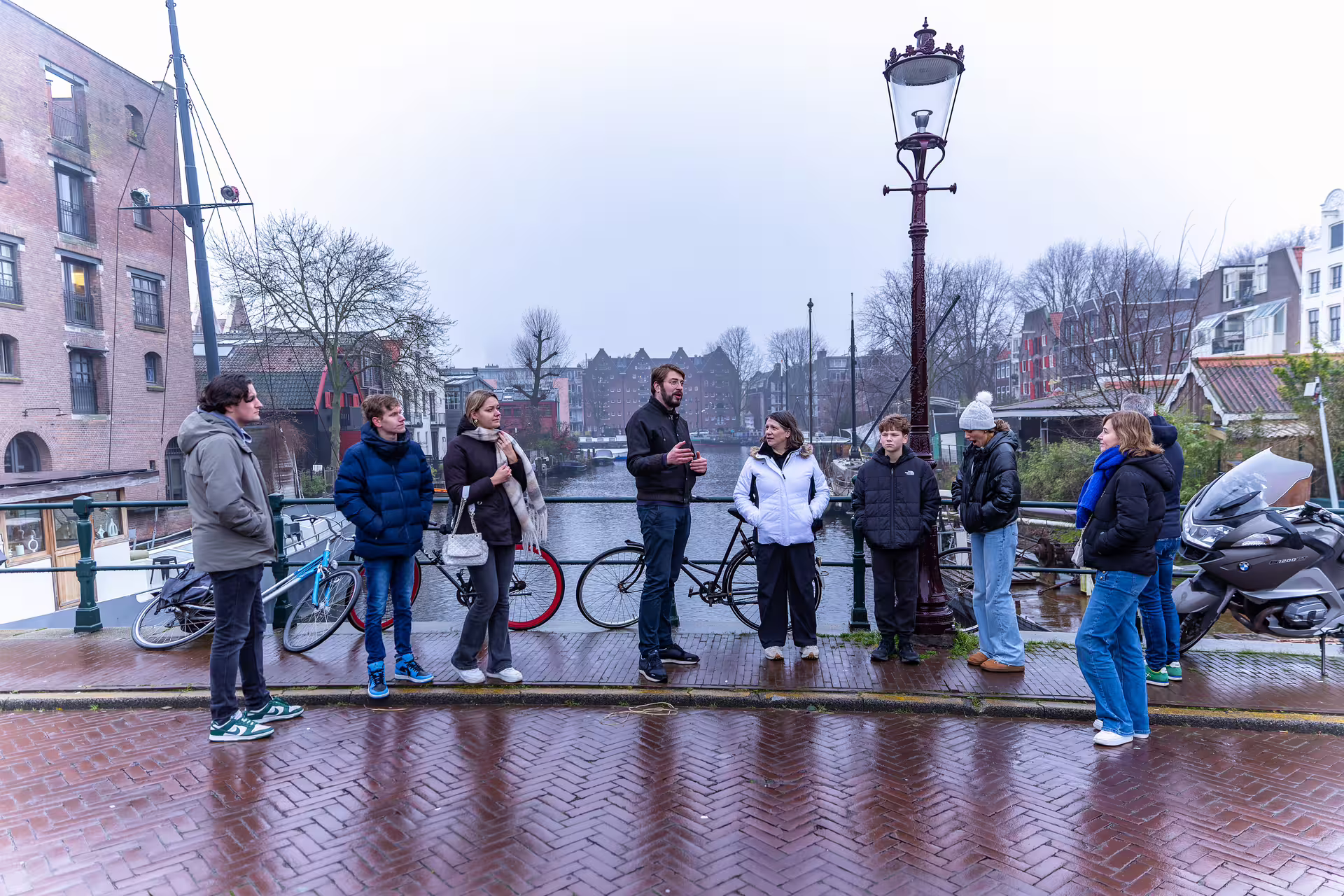 A group of people listening to a tour guide standing on a bridge near a canal, with bicycles, buildings, and trees visible in the background