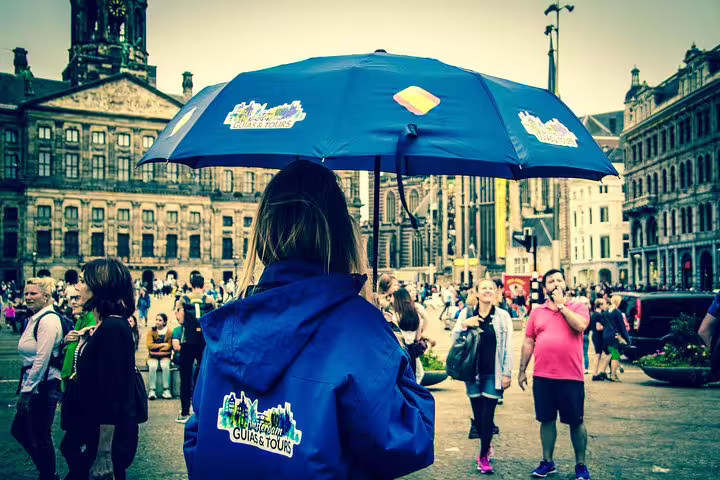 Tour guide in blue jacket and umbrella leading an Amsterdam walking tour near historic buildings in a bustling square.