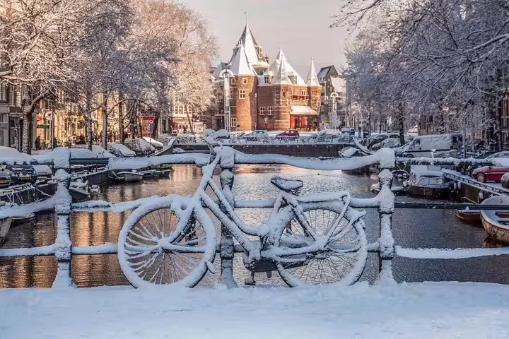 Snow-covered bicycle on an Amsterdam canal bridge with wintry trees and historic architecture, perfect for a winter walking tour.