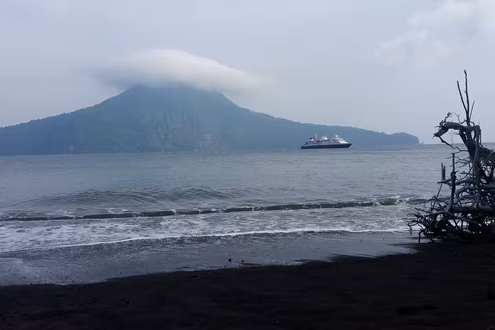 View of Anak Krakatau volcano from black sand beach with boat offshore on Krakatau (Krakatoa) day tour